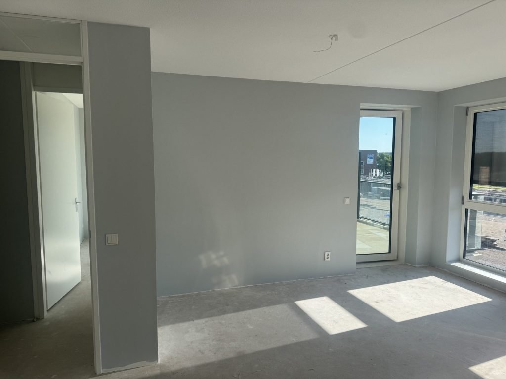 Appartement in aanbouw met een lichtgrijs gesausde wand, een balkondeur en uitzicht op een bouwterrein